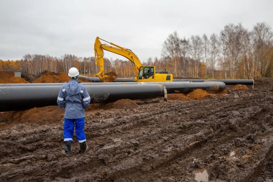 Worker in hard hat stands in front of oil pipeline under construction with yellow heavy equipment.